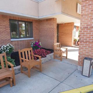 A small outdoor seating area with wooden chairs and flower beds next to the entrance of a brick building.