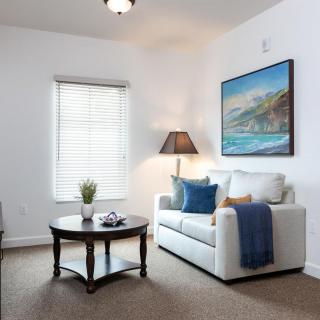 A cozy living room with a white sofa, a round coffee table, and a floor lamp, featuring artwork on the wall and natural light coming through the window.
