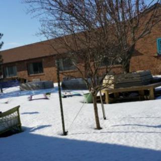 A snowy outdoor area with a bench, a tree, and a brick building in the background.