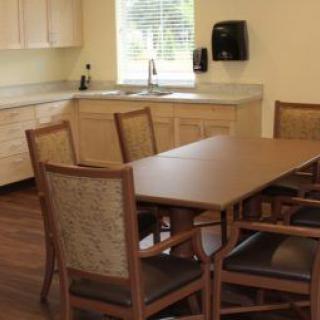 A clean and simple dining area with a table and chairs, adjacent to a kitchen, featuring wooden floors and natural lighting from a window.