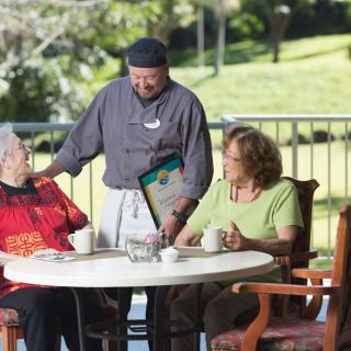 A waiter attending to two elderly women dining outdoors on a sunny day.