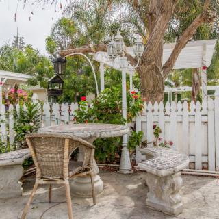 A charming garden patio with a wicker chair, table, white picket fence, and lush greenery.