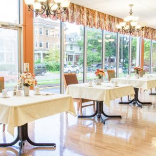 A bright dining room with neatly arranged tables and chairs, featuring large windows that allow natural light to flood the space.