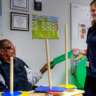 A young woman assists an elderly man during a therapy session in a rehabilitation setting.