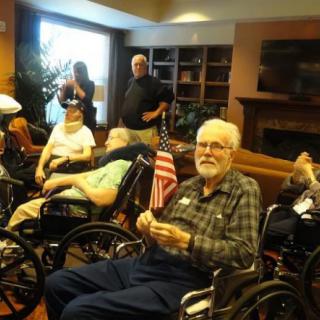 A group of elderly individuals in wheelchairs gathered indoors, with one holding an American flag.