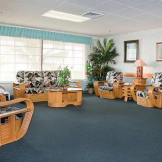 A spacious living room with wooden furniture and floral-patterned upholstery, featuring large windows and indoor plants.