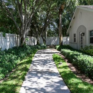 A serene garden pathway leading to a house, surrounded by lush greenery and a white fence.