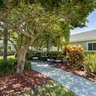 A serene garden with a pathway leading to outdoor seating, surrounded by trees and shrubs on a sunny day.