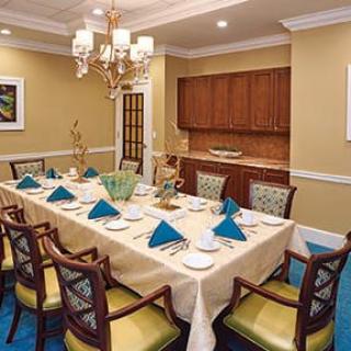 An elegant dining room with a large table set for a formal meal, featuring blue napkins, cushioned chairs, and a chandelier overhead.