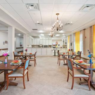 A well-lit dining area with elegantly arranged tables and chairs, featuring a chandelier and decorative flowers.