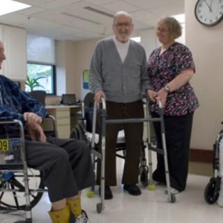 Elderly individuals in a nursing home setting, with one person using a walker and others seated in wheelchairs, accompanied by a caregiver.