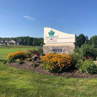 A welcoming sign surrounded by vibrant flowers and greenery, set against a clear blue sky.