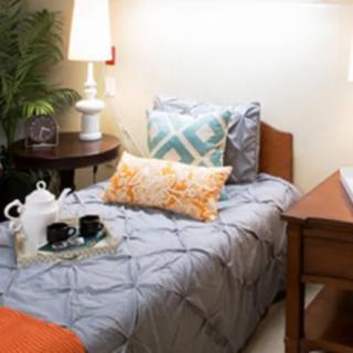 A cozy bedroom with a neatly made single bed, accentuated by decorative pillows and a warm table lamp, complemented by a tea set on the nightstand.