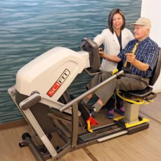 A senior citizen receiving guidance from a trainer while using exercise equipment in a rehabilitation setting.