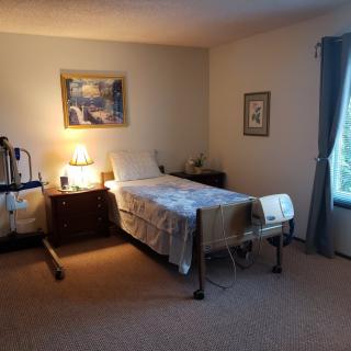 A cozy bedroom with a hospital bed, warmly lit by a lamp on a nightstand, and decorated with a painting on the wall.