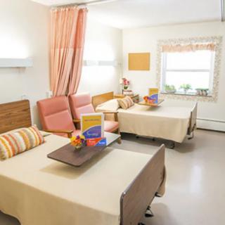 A well-lit hospital room featuring two patient beds with bedside tables, chairs, and a window view.