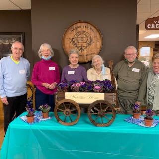 A group of seniors smiling and standing behind a decorated table at an event.