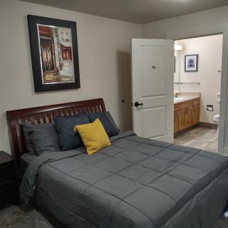 A cozy bedroom with a neatly made bed, featuring gray bedding and a yellow accent pillow. An open door leads to an adjoining bathroom.
