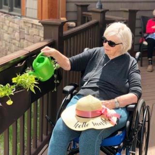 An elderly person in a wheelchair is watering plants outside, wearing sunglasses and holding a sun hat.