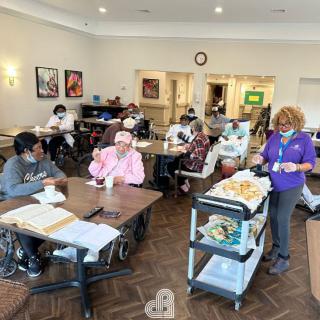 Residents socializing in a nursing home dining area with a healthcare worker serving meals.