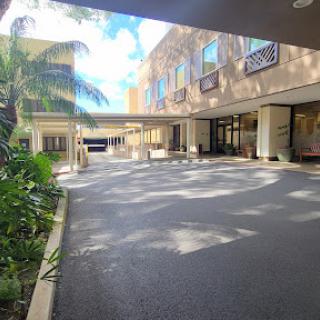 A serene courtyard view of a building, featuring palm trees and a spacious parking area illuminated by sunlight.