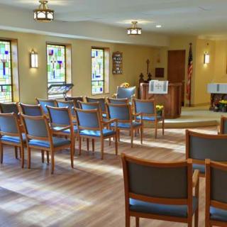 A small chapel with rows of chairs facing a modest altar, surrounded by stained glass windows.