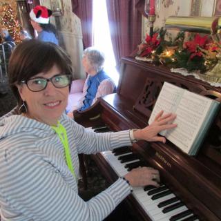 A woman wearing glasses plays the piano in a room decorated for the holidays.