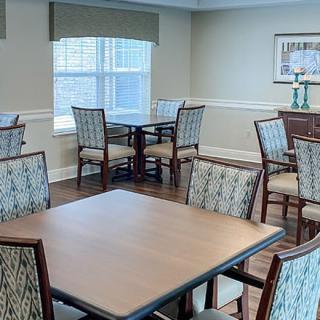A well-lit dining area with multiple tables and chairs, featuring a window and hardwood flooring.
