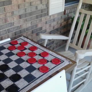 A checkers game set up on a table next to a rocking chair, positioned outdoors against a brick wall.