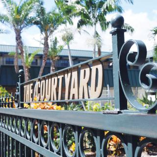 A decorative metal fence with a sign reading 'The Courtyard', set against a backdrop of palm trees and buildings.