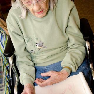 An elderly woman seated in a wheelchair, looking down at a piece of paper with a focused expression.
