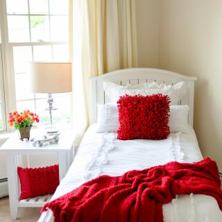 A cozy bedroom with white bedding and vibrant red accents, including a pillow and throw blanket. Natural light streams in through the window, illuminating the room.
