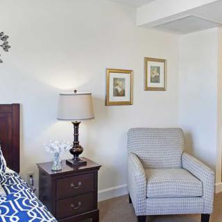A cozy bedroom corner featuring a patterned chair, a nightstand with a lamp, and framed artwork on the wall.