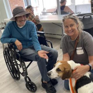 A man in a wheelchair interacts with a therapy dog and its handler indoors.