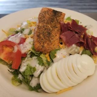 A fresh salad topped with salmon, bacon, sliced boiled eggs, tomatoes, lettuce, and cheese drizzled with dressing.