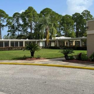 A low-rise building surrounded by lush greenery and palm trees, with a neat driveway in the foreground.
