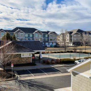 A residential apartment complex with a view of a parking lot and tennis court under a partly cloudy sky.