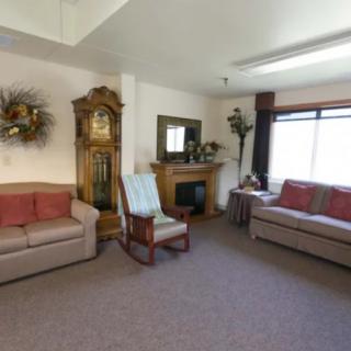 A cozy living room featuring sofas, a rocking chair, a grandfather clock, and a fireplace with a mirror above.