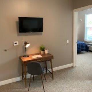 A cozy study area with a desk, chair, and lamp next to a wall-mounted TV, leading into a bedroom.