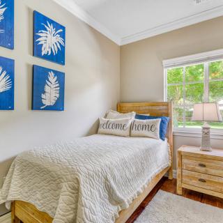 A cozy bedroom with a single bed and decorative pillows, featuring blue leaf-themed wall art and a wooden dresser by the window.