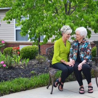 Two elderly women sitting on a bench and smiling in a garden setting.