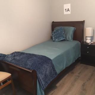 A neatly made single bed in a small bedroom with blue bedding and a nightstand with a lamp.