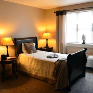 A cozy bedroom with a single bed, warm lighting from lamps, and a large window with curtains.