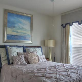 A cozy bedroom with a neatly made bed, featuring decorative pillows and a large painting above the headboard. A lamp stands beside the bed near a window with drapes.