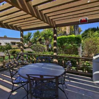 A sunny outdoor patio with a pergola, featuring a table and chairs, surrounded by lush garden landscaping.
