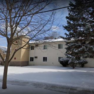 A two-story residential building covered in snow, surrounded by trees under a cloudy sky.