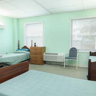 A bright and simple bedroom featuring two beds, a chair, and a dresser, with light green walls providing a calming atmosphere.