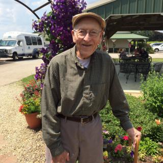 An elderly man wearing a hat and glasses stands outdoors near plants.
