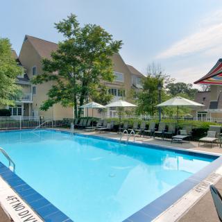 A residential outdoor swimming pool with lounge chairs and umbrellas, surrounded by trees and buildings.