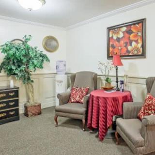 A cozy living room corner with two armchairs, a small table covered with a red cloth, and decorative elements including a lamp, wall art, and an indoor plant.
